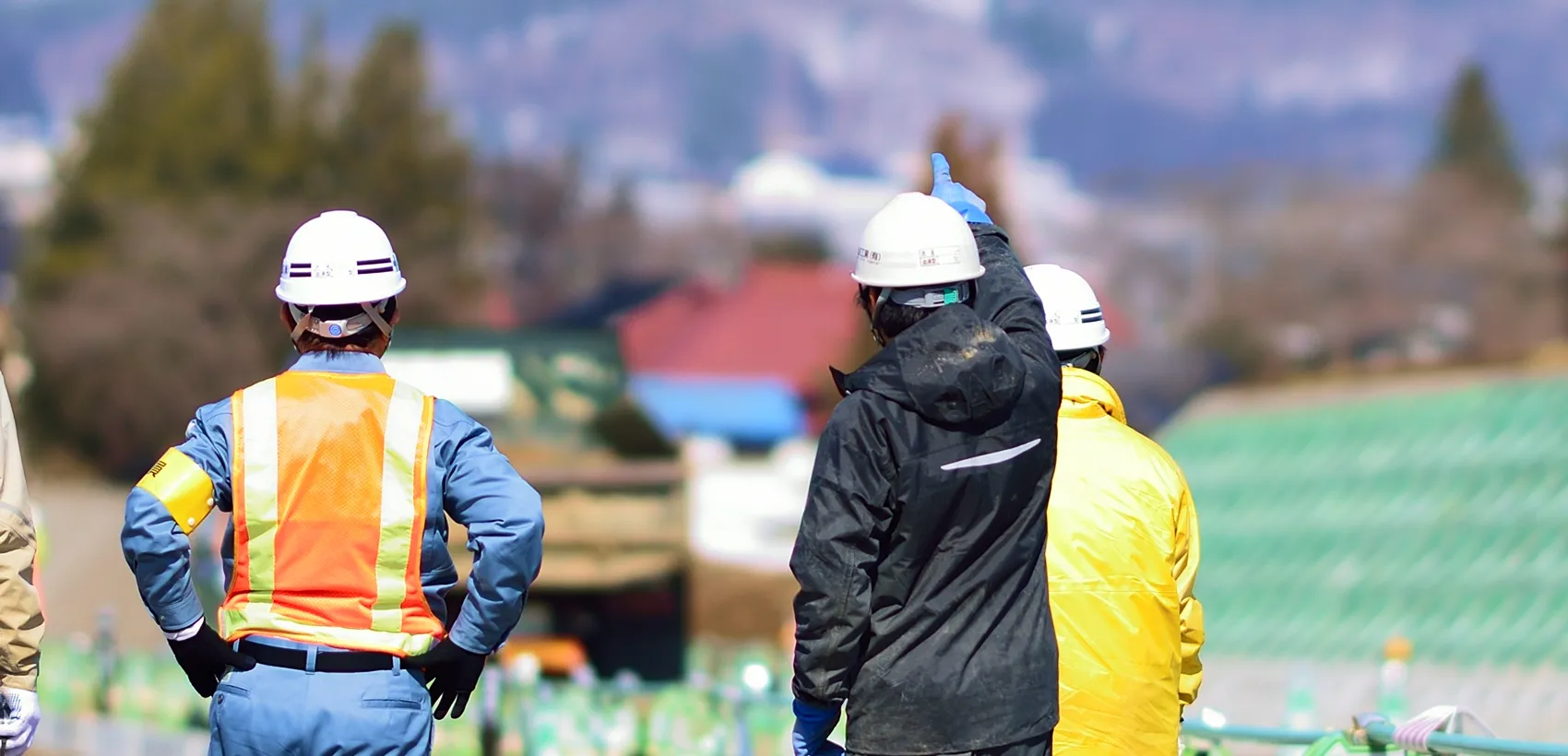 建設現場で白ヘルメットを着用した作業員たちが後ろ姿で立ち、遠くの現場を指差して確認している様子