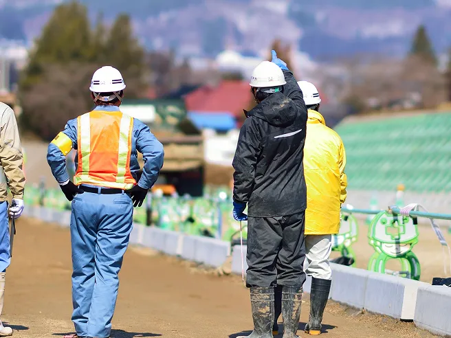 建設現場で白ヘルメットを着用した作業員たちが後ろ姿で立ち、遠くの現場を指差して確認している様子